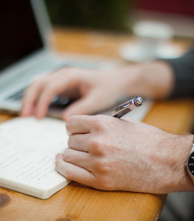 Male hands taking notes in notebook on a desk with laptop open, only hand, pen and wristwatch in focus