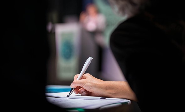 Woman's hand taking notes with white pen during a speech, only hand and pen in focus, rest of picture blurry