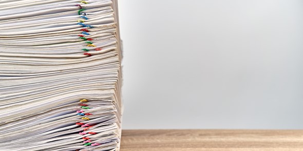 Large stack of files with multicoloured paperclips attached on a wooden desk, stack aligned to the left of the picture