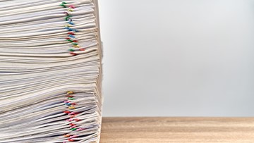 Large stack of files with multicoloured paperclips attached on a wooden desk, stack aligned to the left of the picture